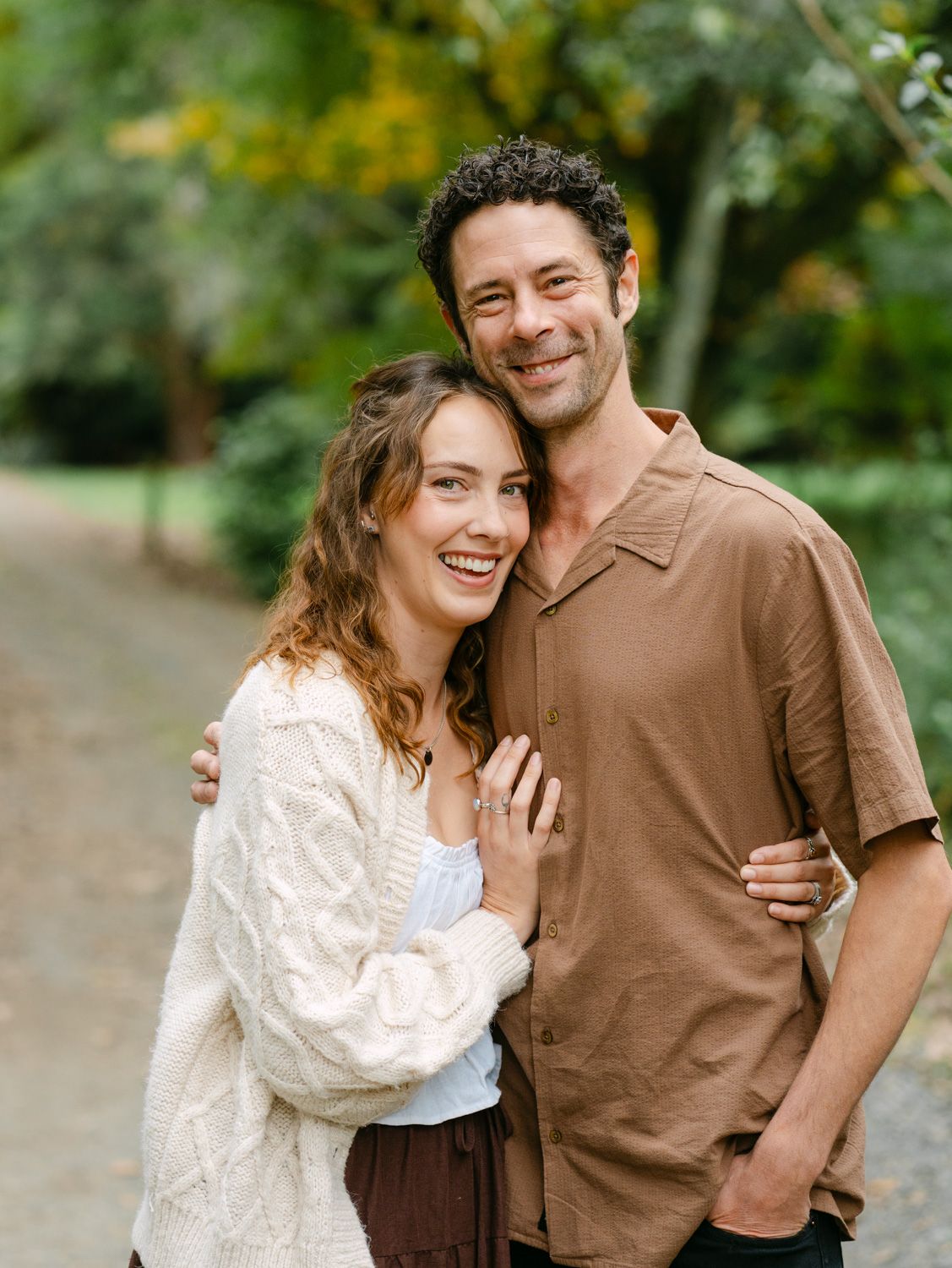 A smiling couple embraces outdoors on a tree-lined path, woman in white knit cardigan and man in brown shirt.