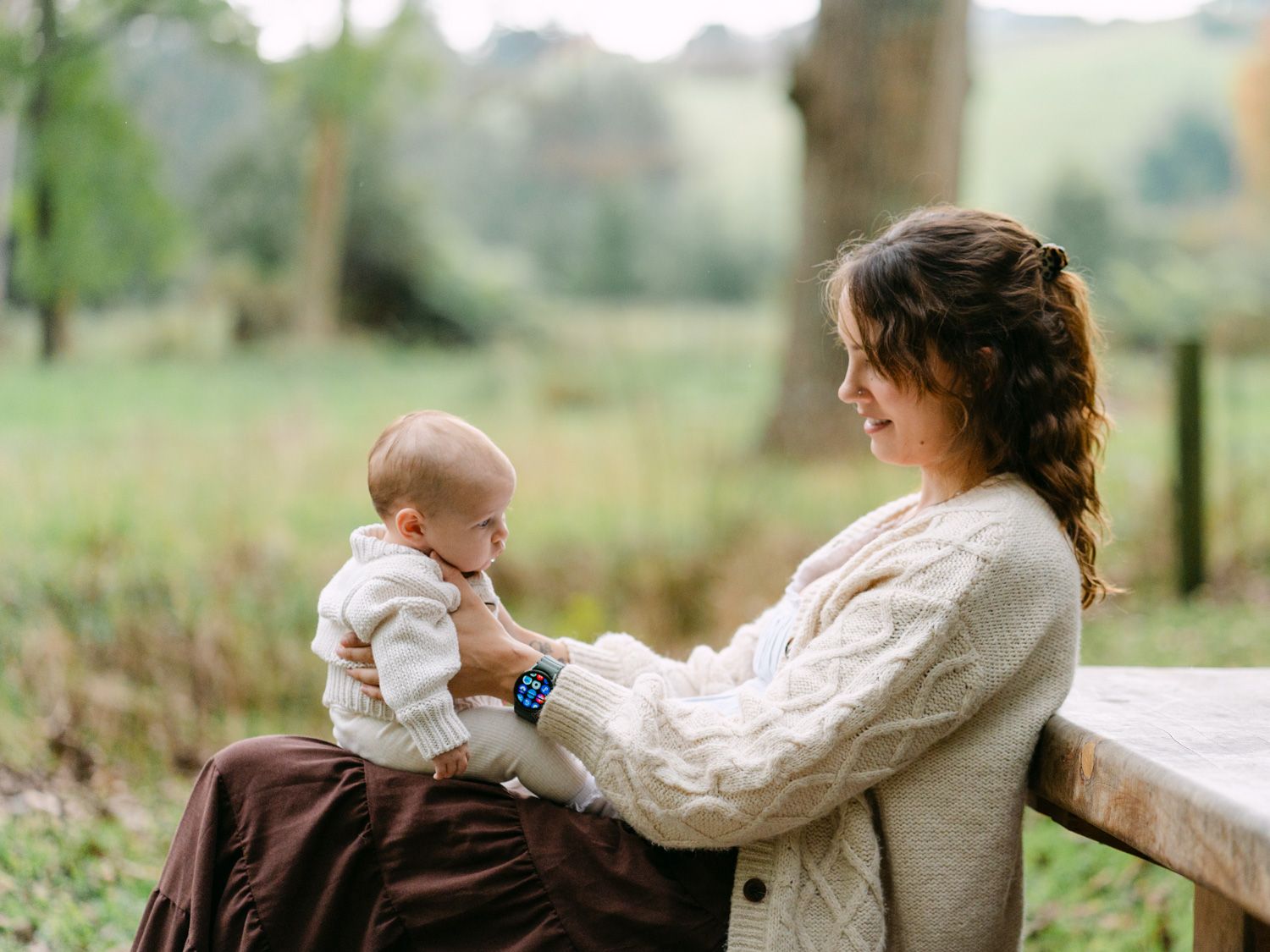 A mother sits on a wooden bench outdoors, lovingly holding her baby in a lush green countryside setting.