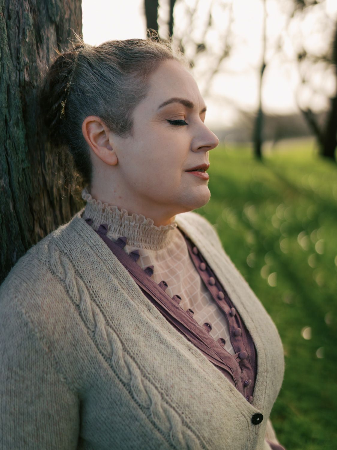 Woman in cream cardigan leaning against tree with eyes closed in golden hour light outdoors.