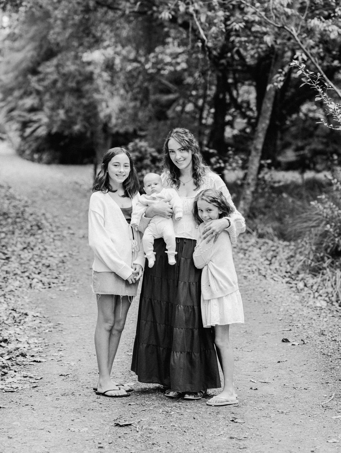 Black and white photo of a mother holding a baby, flanked by two young girls, posing on a wooded path.