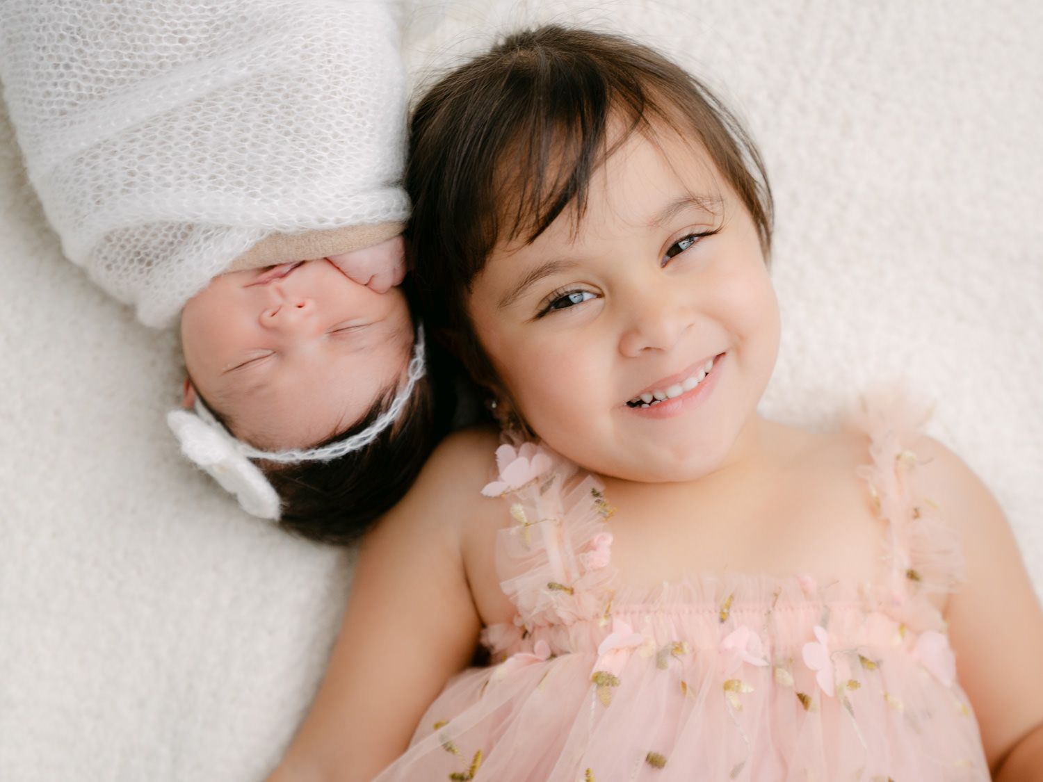 A smiling toddler girl in a pink tutu dress poses with her newborn sibling wrapped in white on a soft white blanket.