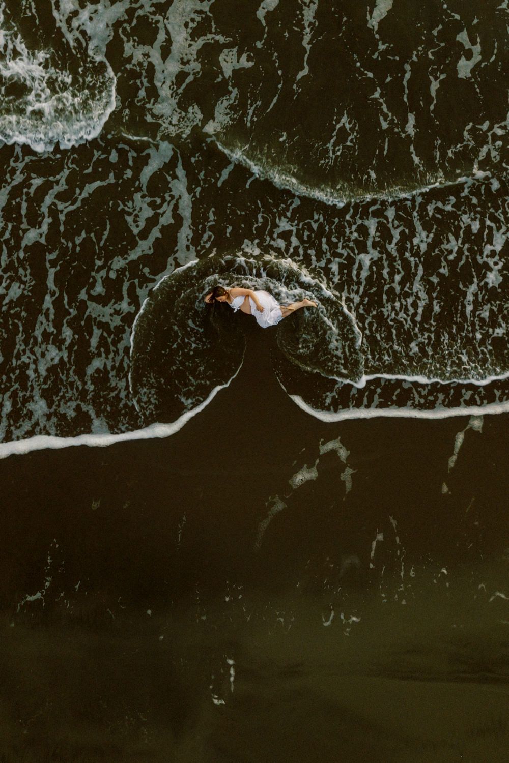 Aerial view of a person lying on dark wet sand as ocean waves wash around them on a moody beach.