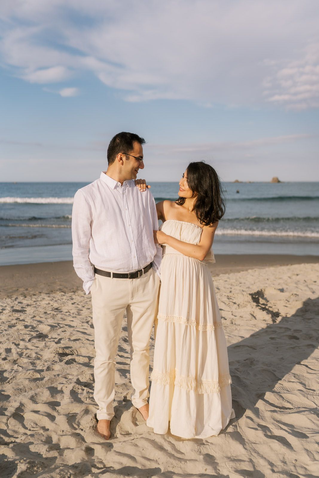 A couple in matching cream outfits share a romantic moment on a sandy beach with ocean waves behind them.