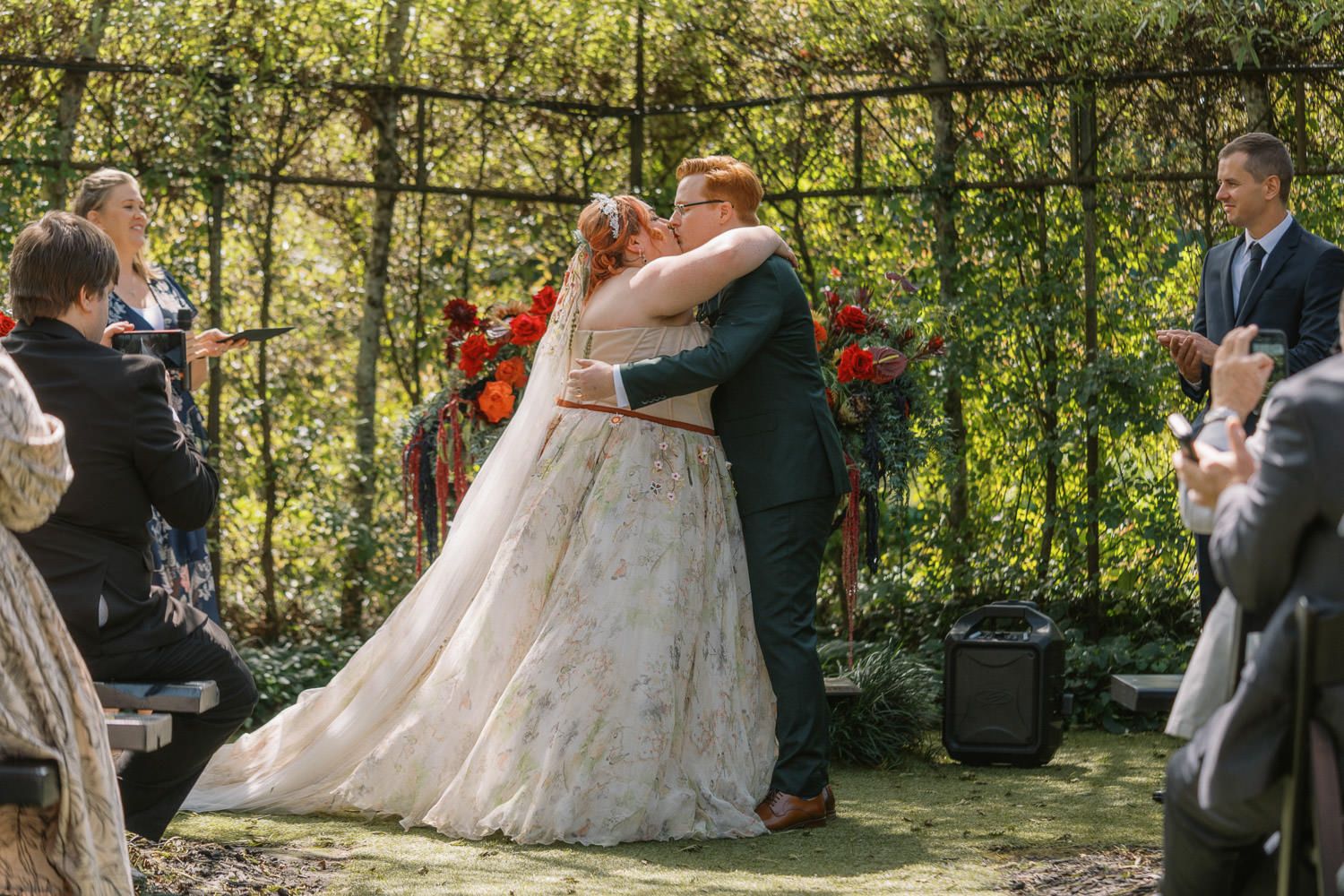 Bride and groom share first kiss at outdoor wedding ceremony surrounded by guests and floral arch backdrop.
