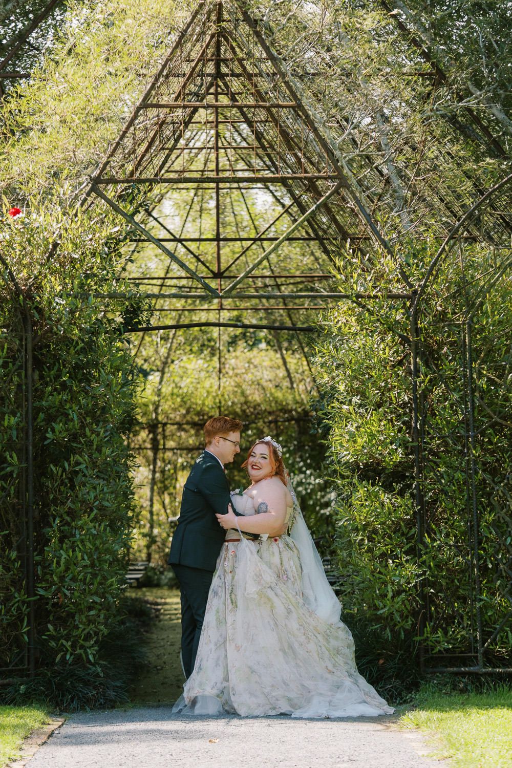 Bride and groom embrace under ornate metal garden arch surrounded by lush greenery on their wedding day.