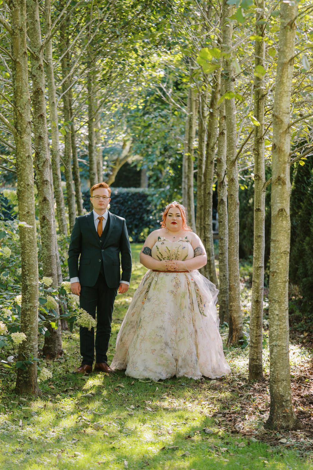 A bride in a floral gown and groom in a dark suit walk among tall slender trees in a lush garden setting.