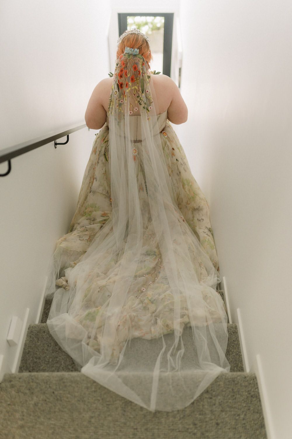 Bride in floral wedding gown with tulle veil walks up carpeted stairs, viewed from behind, hair adorned with flower clip.