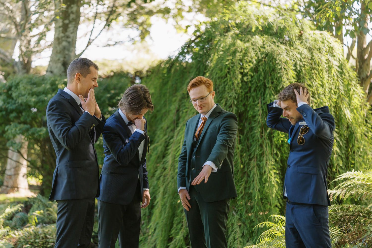 Four groomsmen in dark suits laughing and joking outdoors near a weeping willow tree at a wedding.