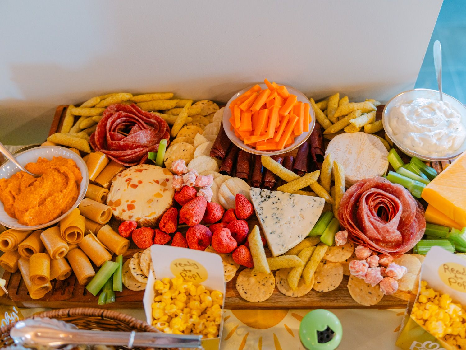 A colorful charcuterie board with meats, cheeses, carrots, strawberries, crackers, and dips arranged on wood.