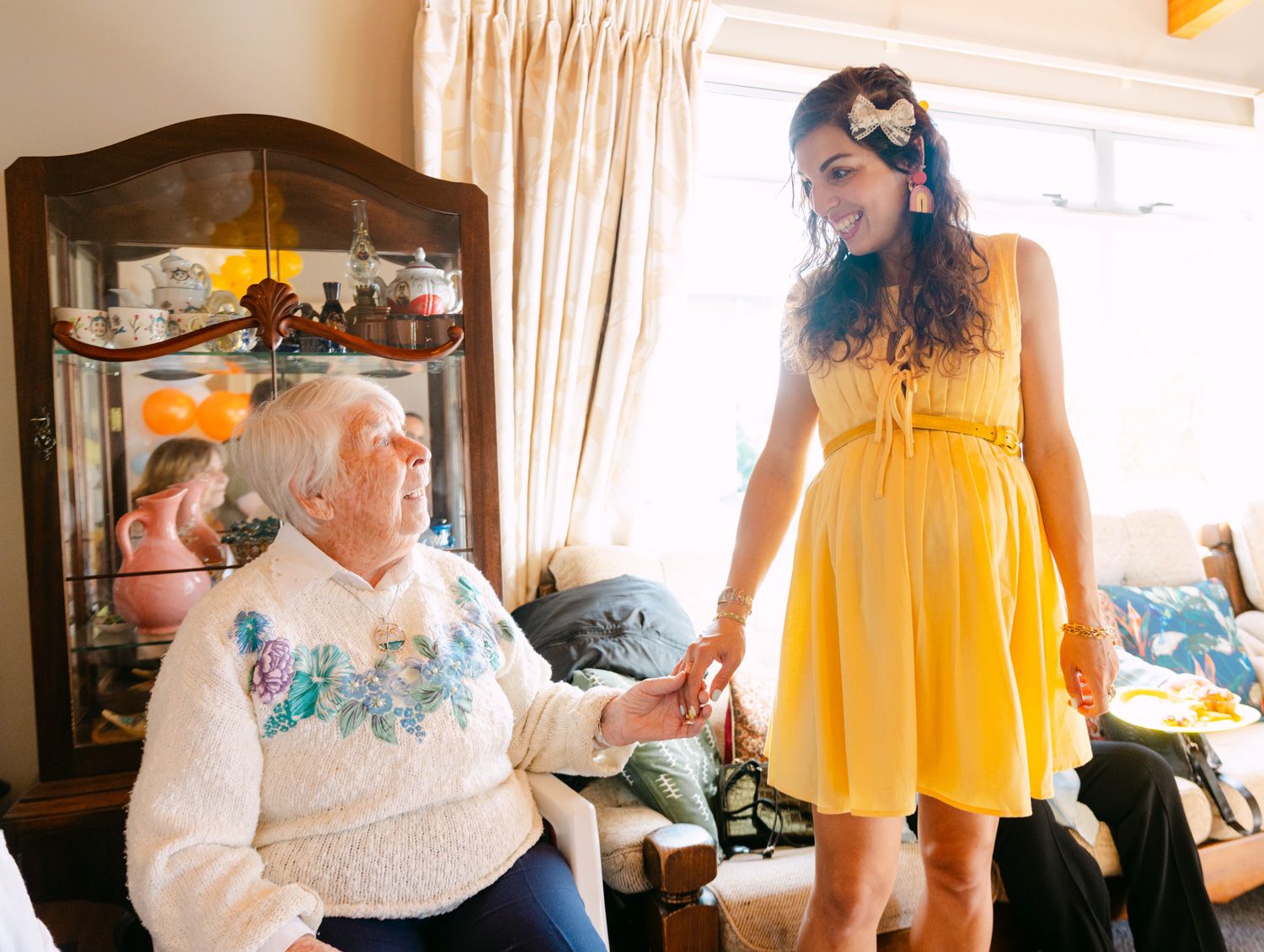Elderly woman in white sweater holds hands with young woman in yellow dress indoors.