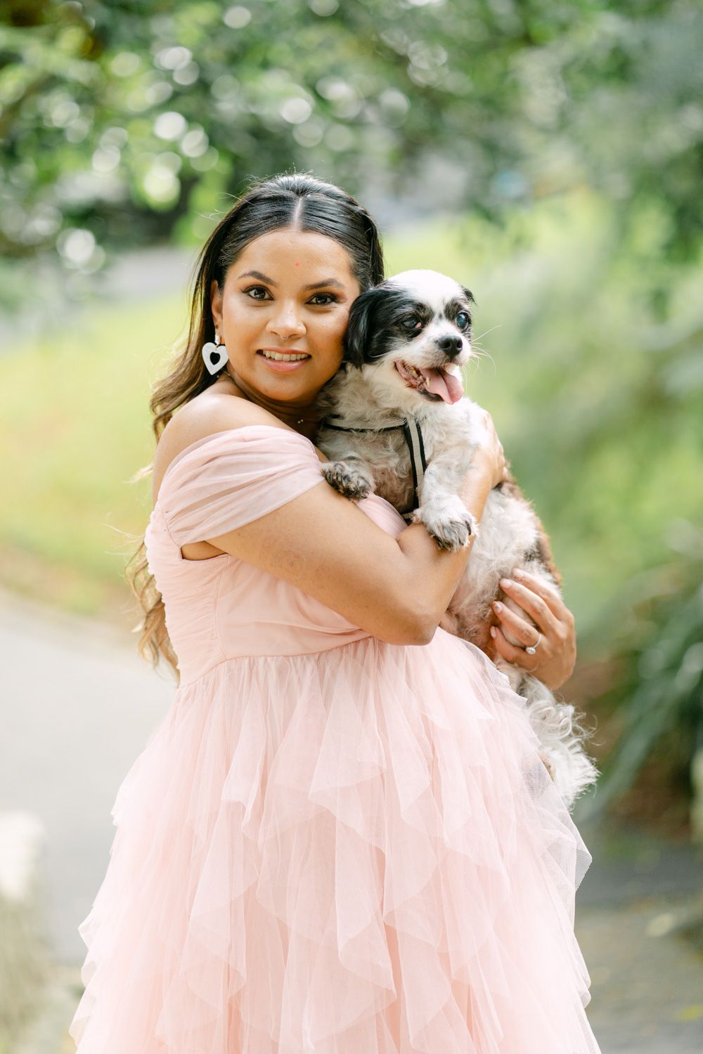 Woman in pink tulle dress holding a small black and white dog outdoors in a lush green garden setting.