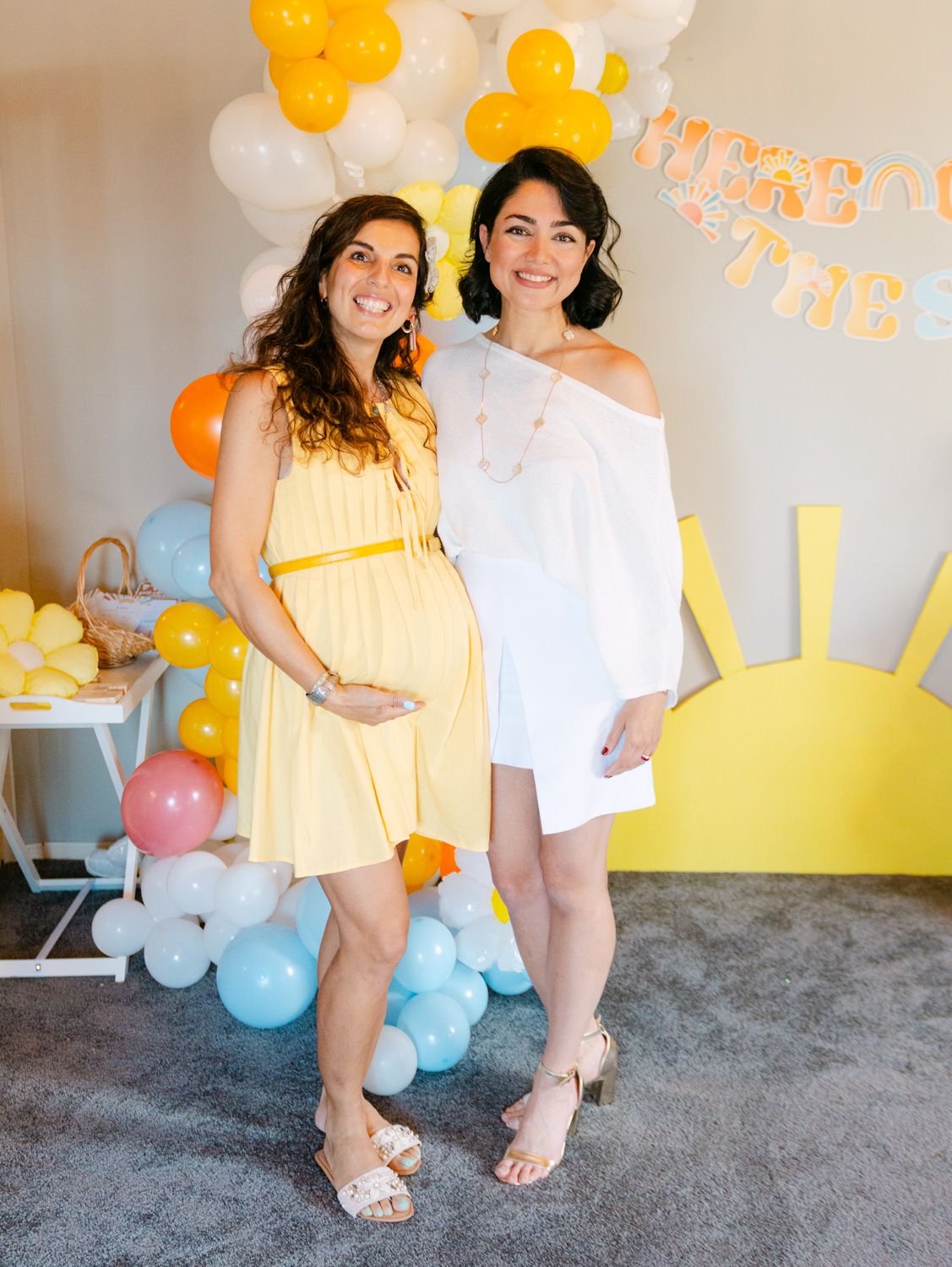 Two women posing at a sunny baby shower with yellow and blue balloon arch decorations.