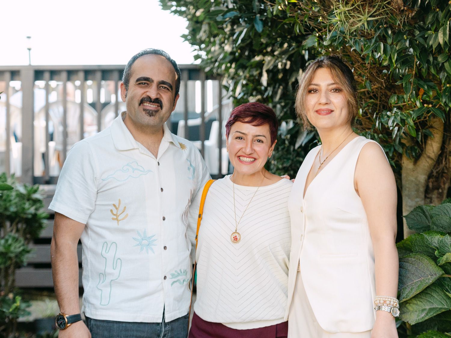 Three smiling adults posing together outdoors near green trees at a casual gathering.