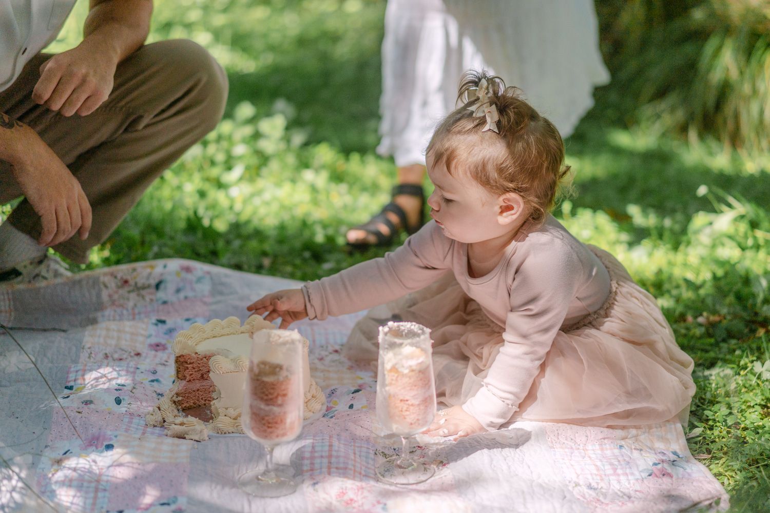 Toddler girl in pink dress reaching for mason jar candles on a blanket in a sunny garden.