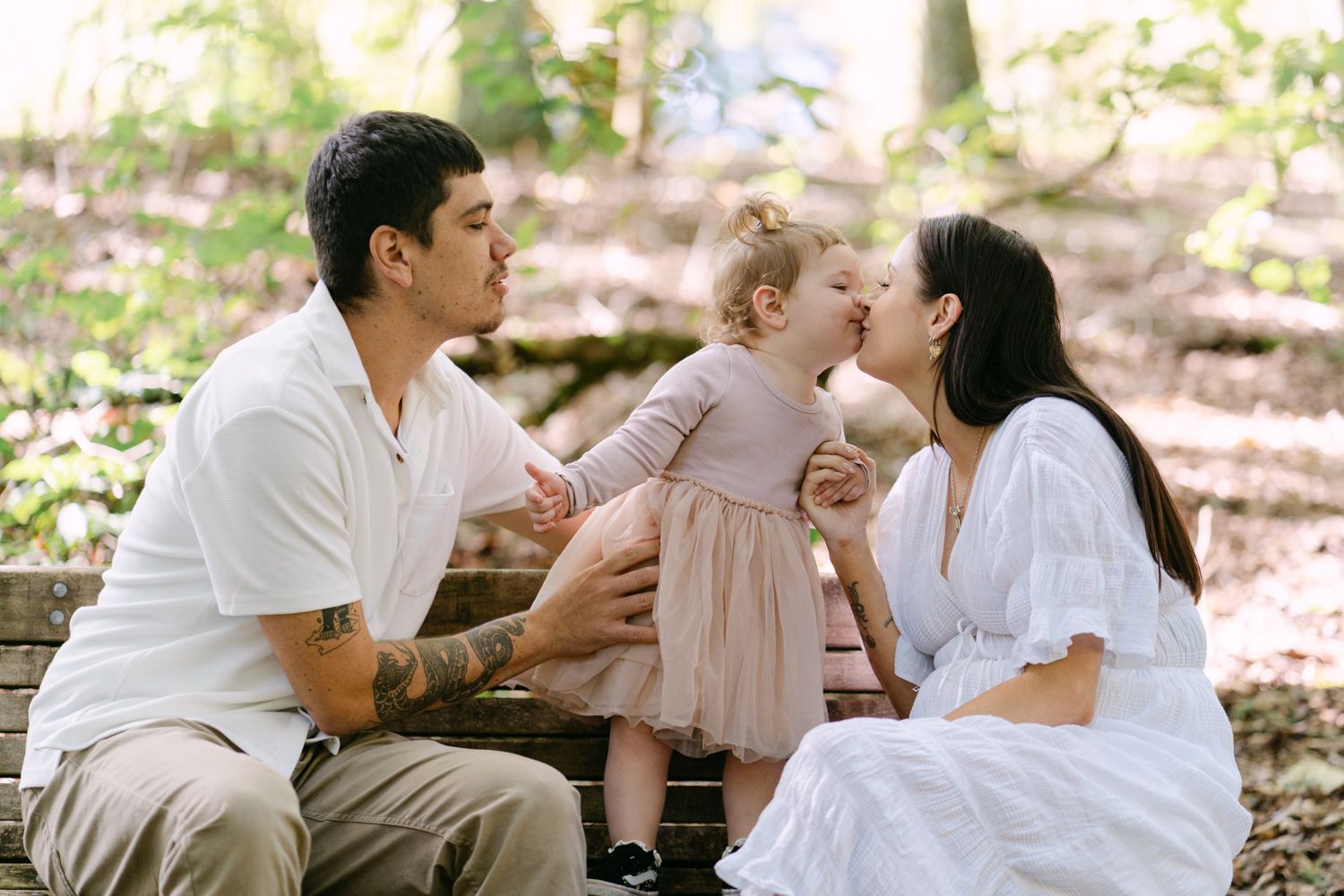 A young family sits on a wooden bench in a park, with parents kissing their toddler daughter in a pink dress.