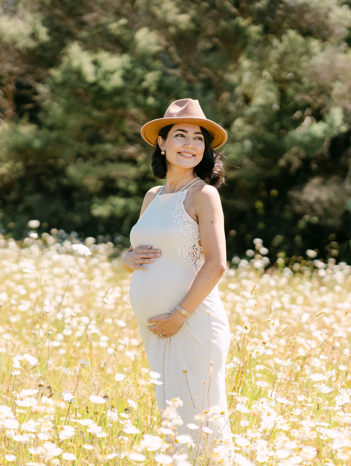 Smiling pregnant woman in white dress and tan hat cradling baby bump in sunny wildflower meadow.
