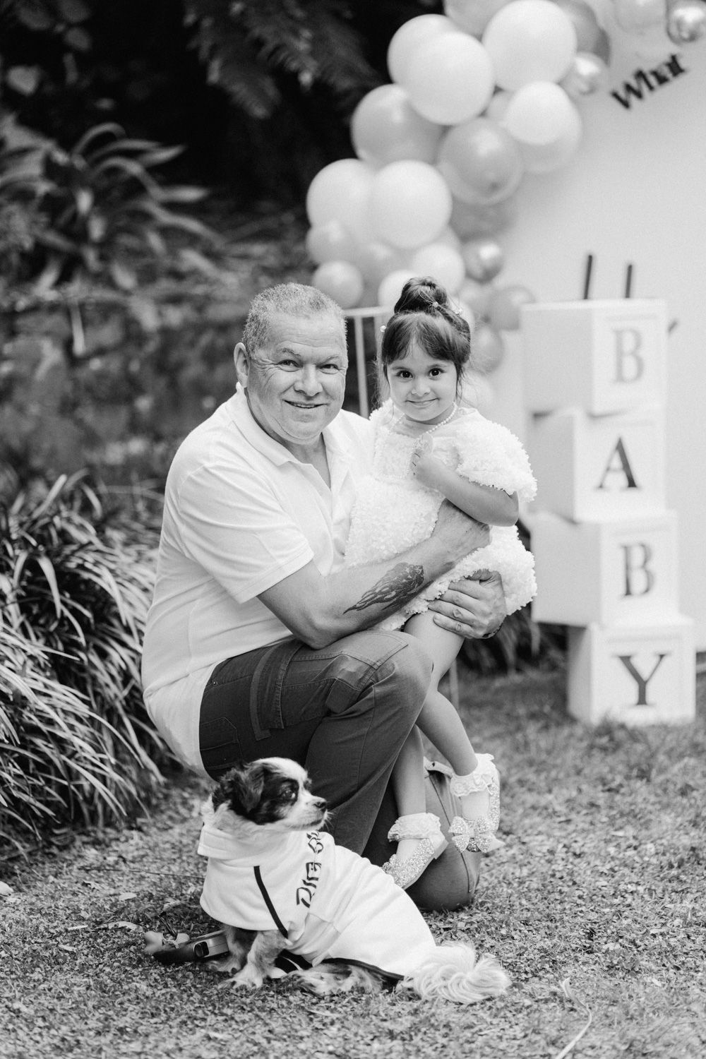 Man crouching with young girl and small dog near BABY blocks and balloon arch at outdoor baby shower.