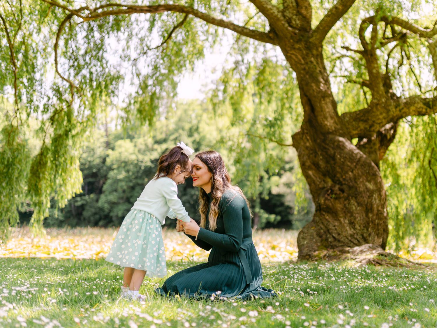 Mother and young daughter sharing a tender nose kiss under a weeping willow tree in a lush green park.