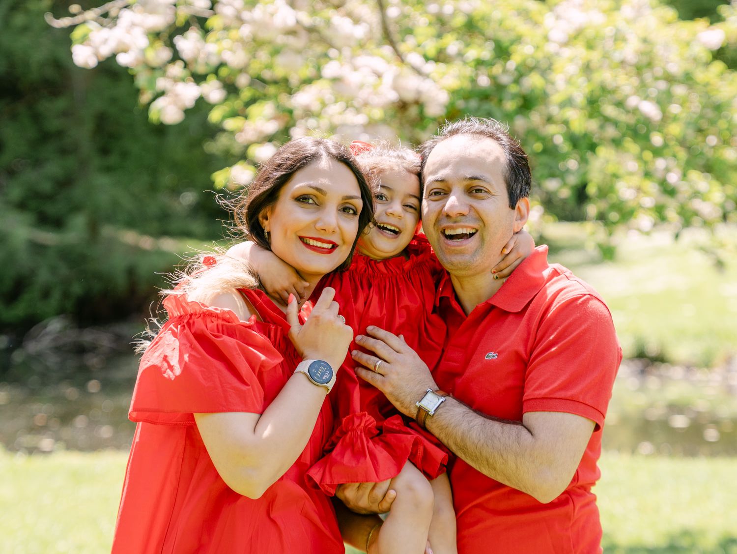 Happy family of three in matching red outfits smiling outdoors with blooming trees in background.