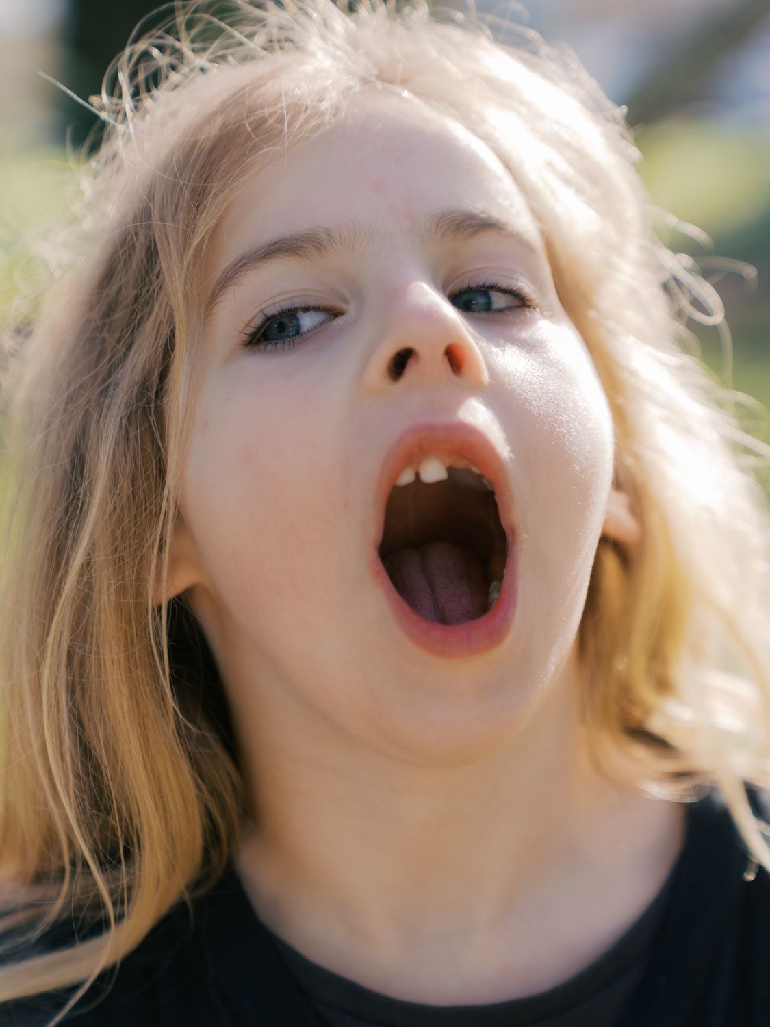 Close-up of a young blonde girl with mouth wide open in a surprised or excited expression outdoors.