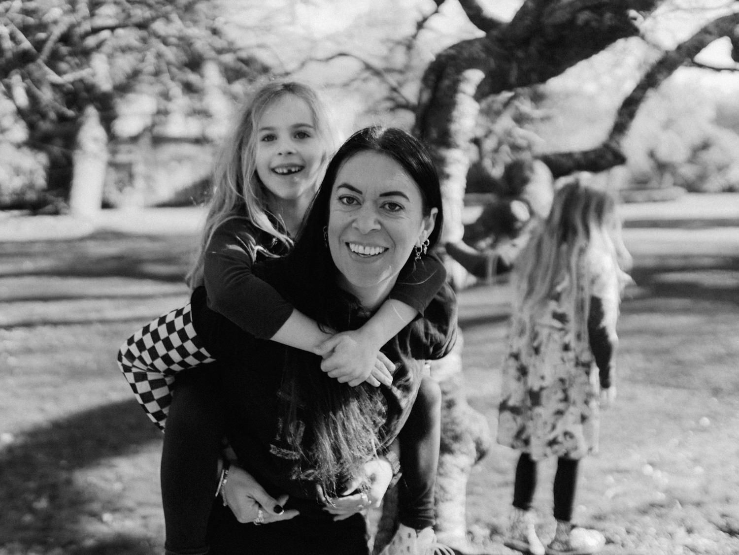 Black and white photo of a smiling woman giving a young girl a piggyback ride in a park.