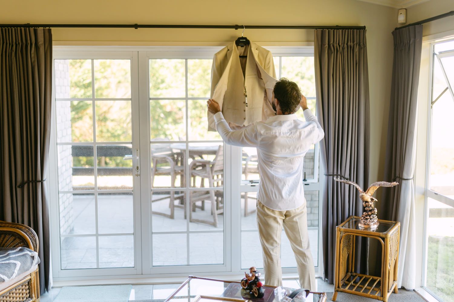 Groom hanging a beige wedding suit jacket on a glass door before getting dressed for his wedding day.