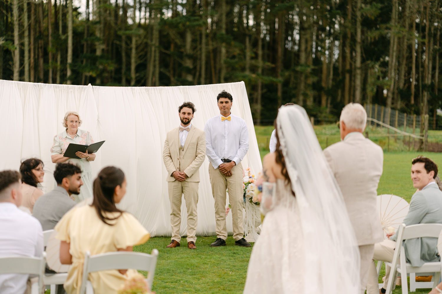 Outdoor wedding ceremony on green lawn with groom awaiting bride, white drape backdrop, surrounded by seated guests.