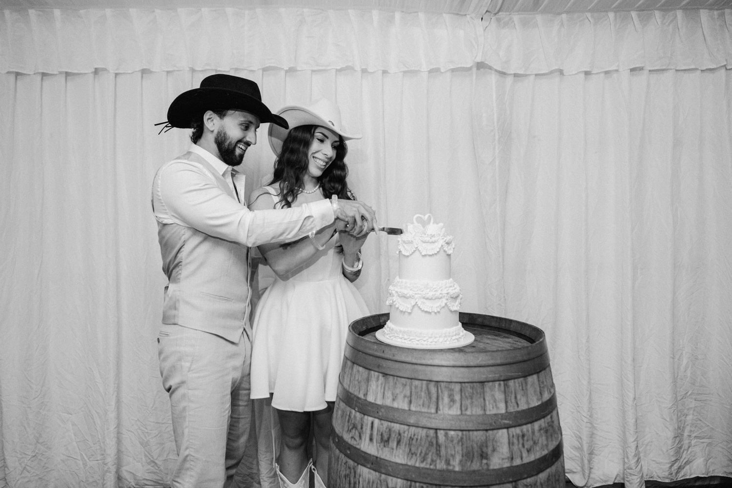 Black and white photo of a couple cutting their wedding cake on a barrel at a rustic country wedding.
