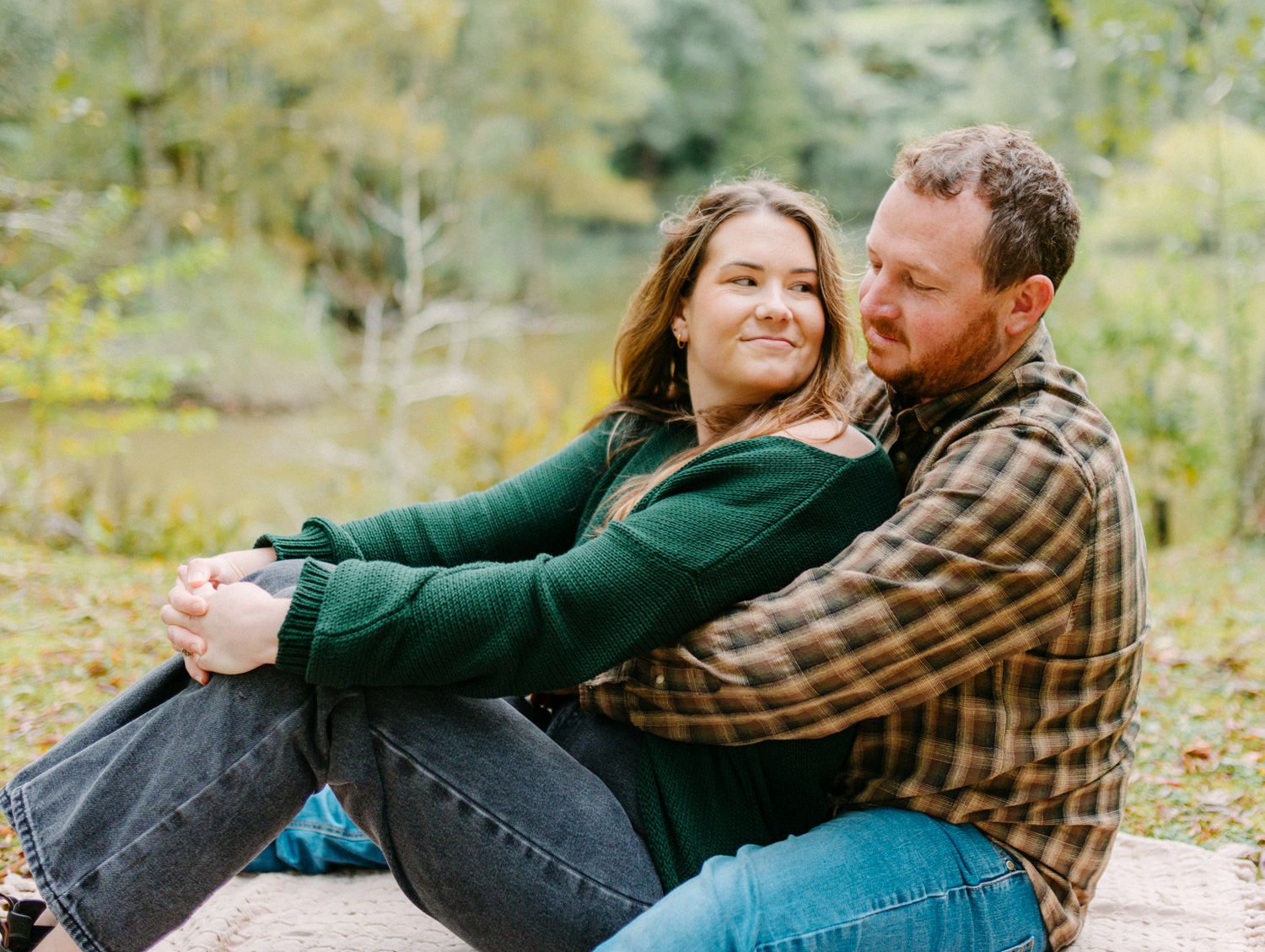 Couple sitting together outdoors in fall, woman in green sweater leaning back against man in plaid shirt, smiling warmly.