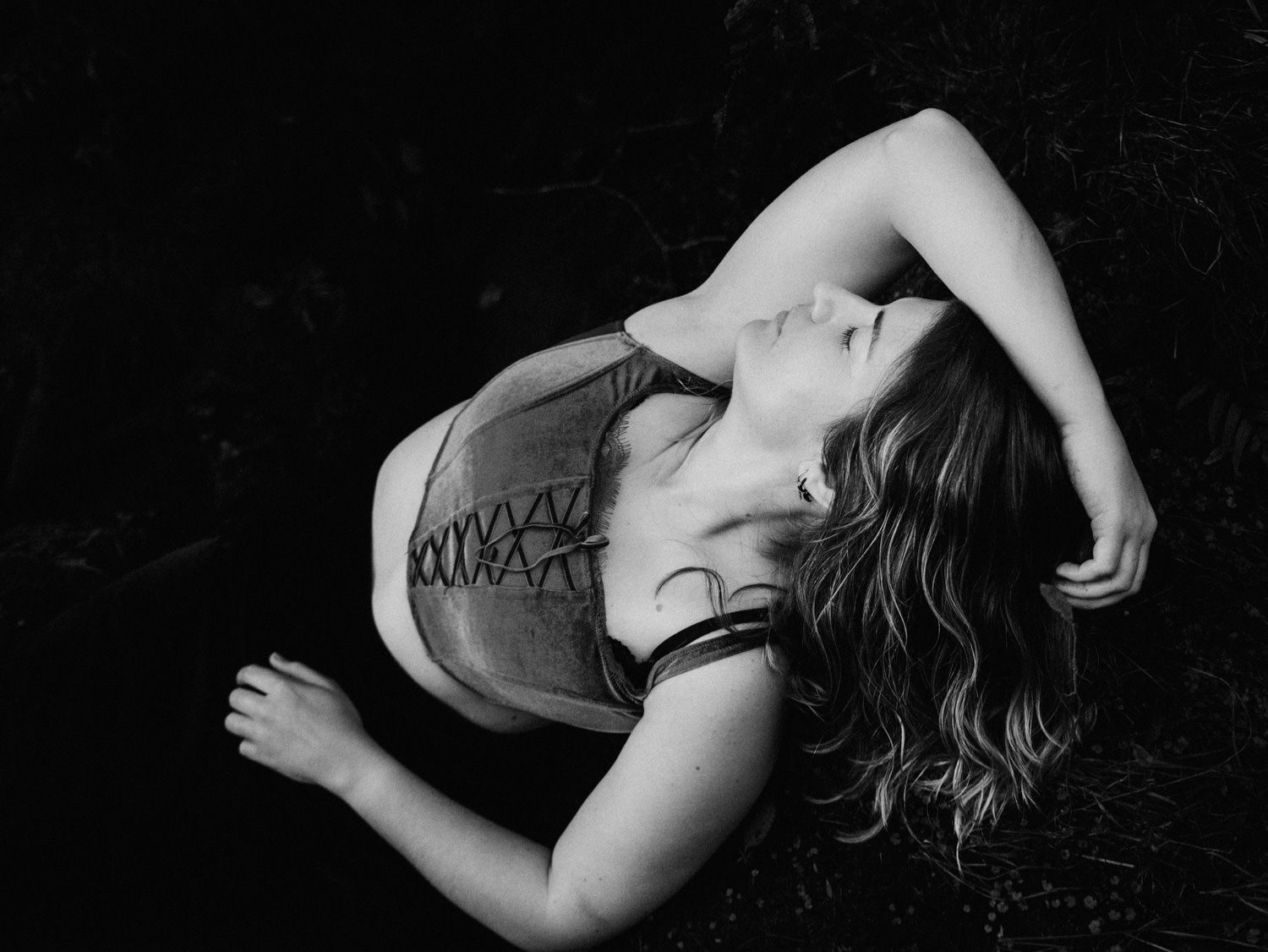 Black and white photo of a woman lying on the ground with eyes closed, wearing a lace top.
