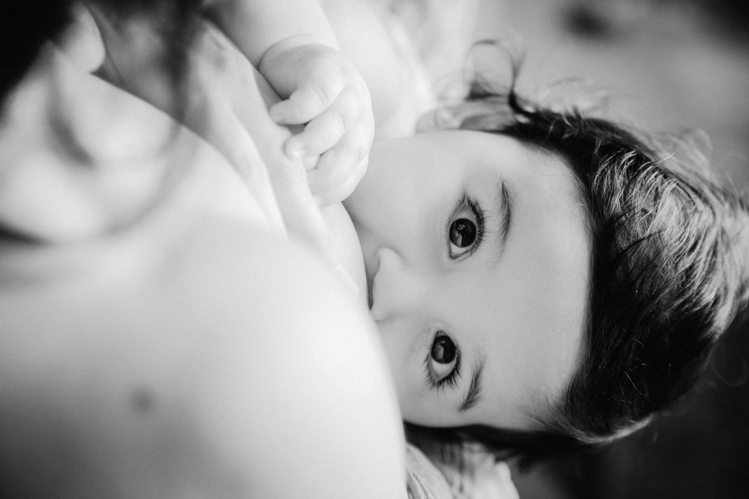 Black and white photo of a baby breastfeeding, gazing up with bright eyes in an intimate moment.
