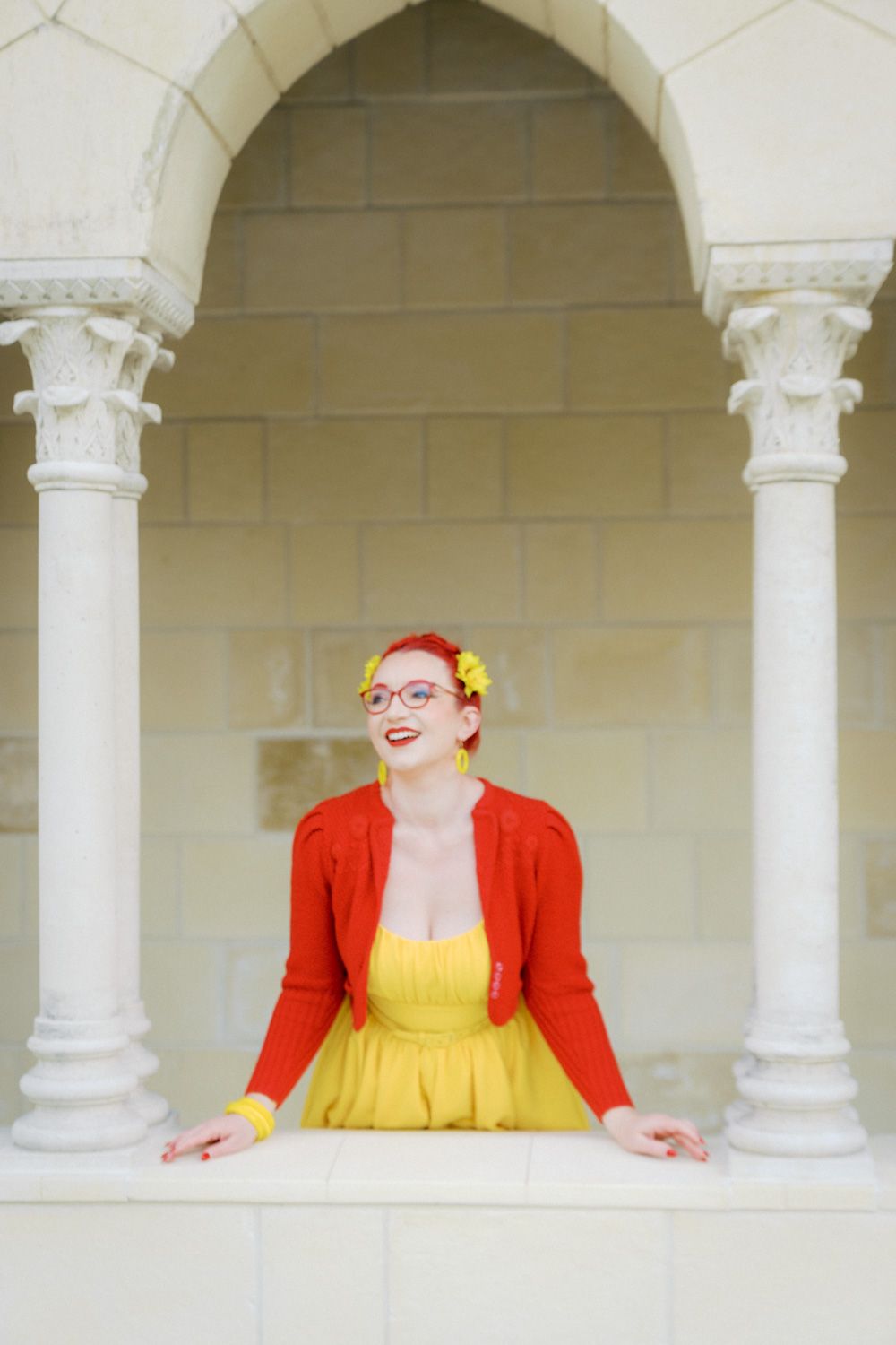 Woman in yellow dress and red cardigan with flower hair accessories poses between white columns of an elegant archway.