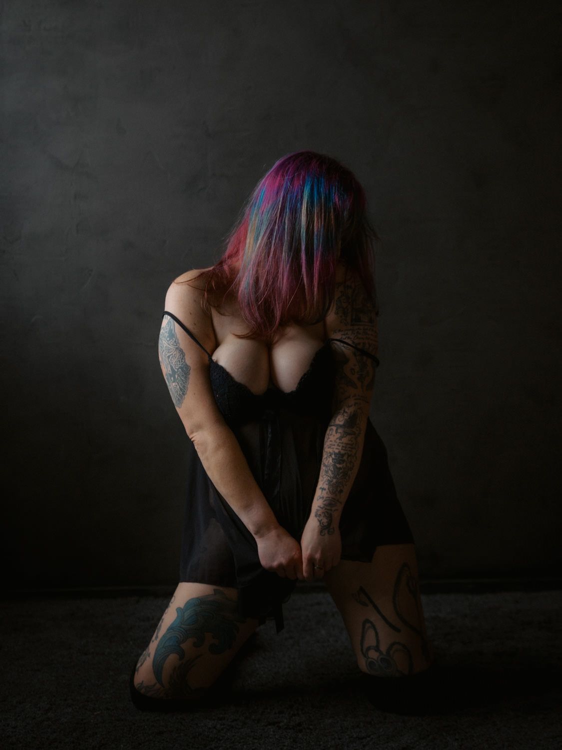 Woman with colorful pink and blue hair, tattoos, kneeling in black dress against dark background.