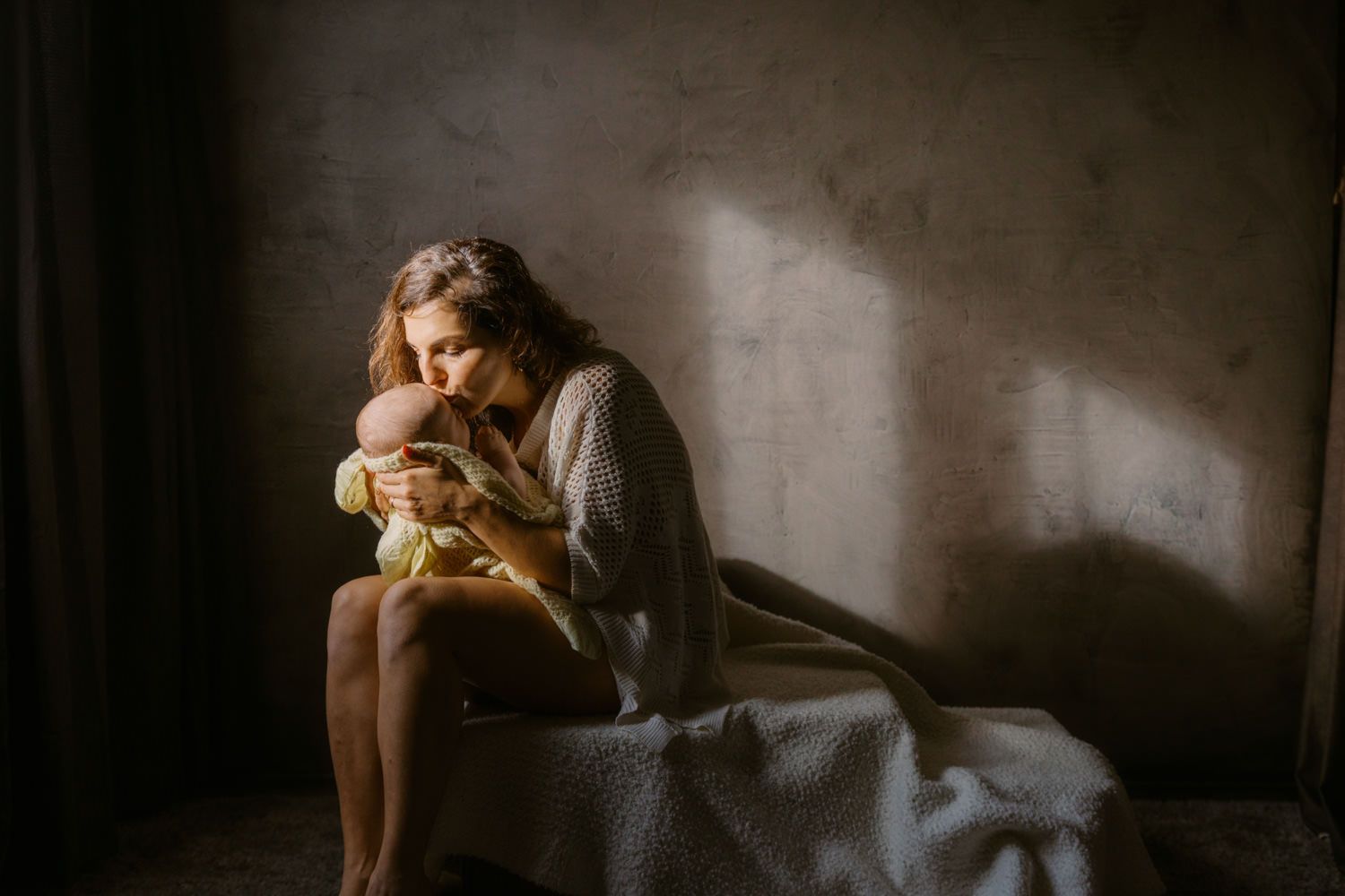 A mother tenderly holds her newborn baby in soft natural light in a serene, moody room.