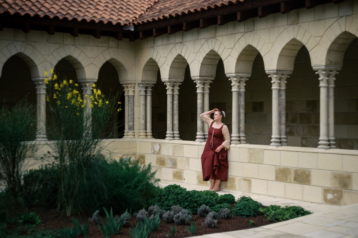 Woman in burgundy dress poses in a medieval stone courtyard with arched colonnade and garden at The Cloisters museum.