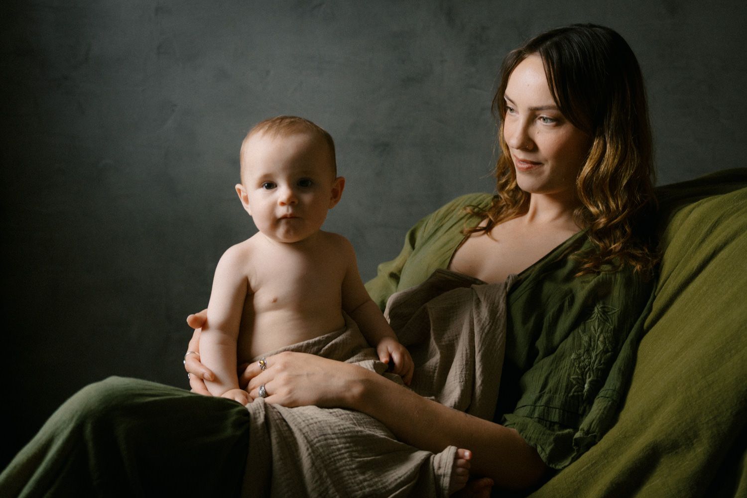 Young mother in green dress sits with curious baby on her lap against dark textured wall.