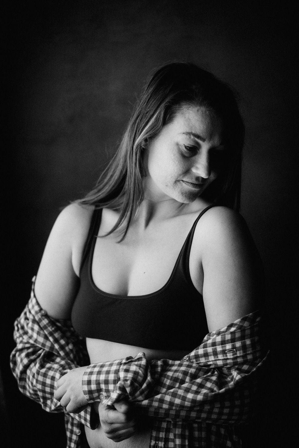Black and white portrait of a young woman in a crop top and plaid shirt looking down in studio lighting.