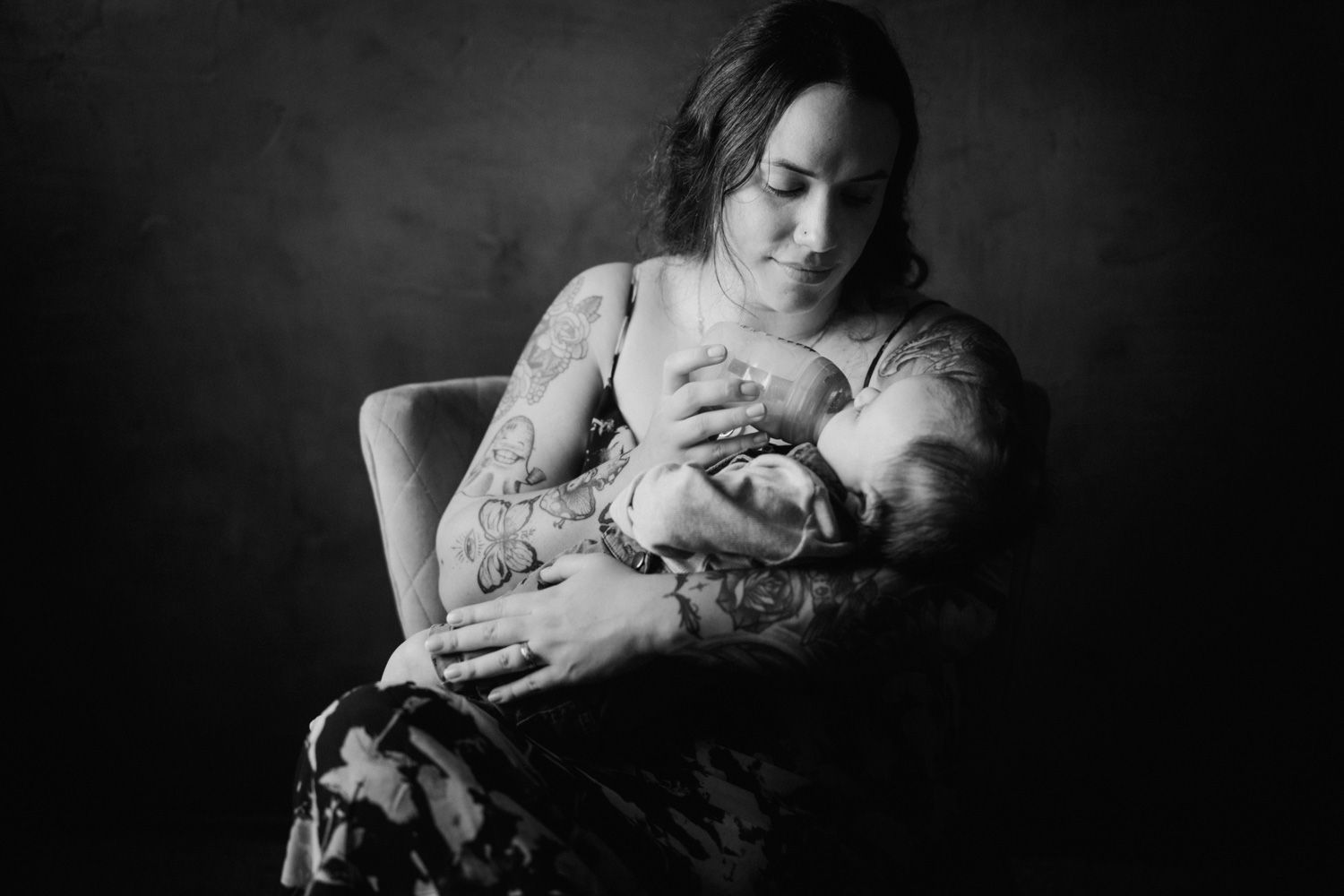 Black and white photo of a tattooed mother tenderly breastfeeding her newborn baby while seated in a chair.