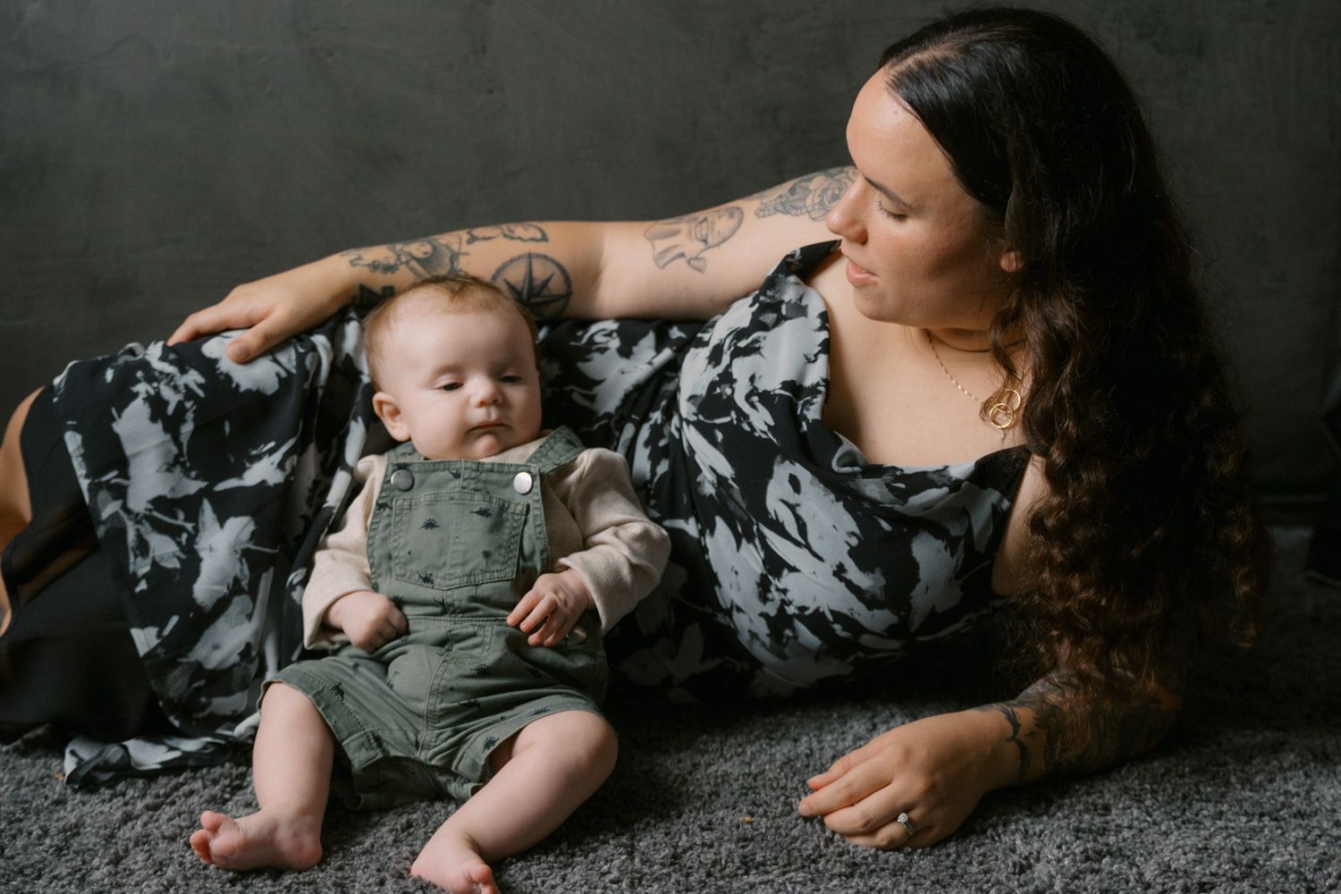 A tattooed mother lovingly gazes at her baby boy in green overalls on a gray carpet.