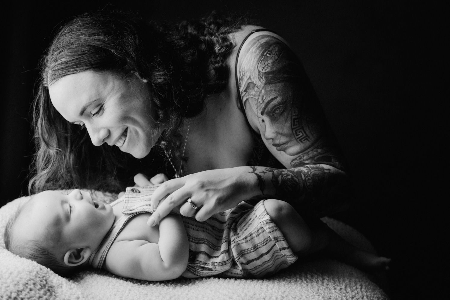 Black and white photo of tattooed mother smiling at her newborn baby lying on a soft blanket.