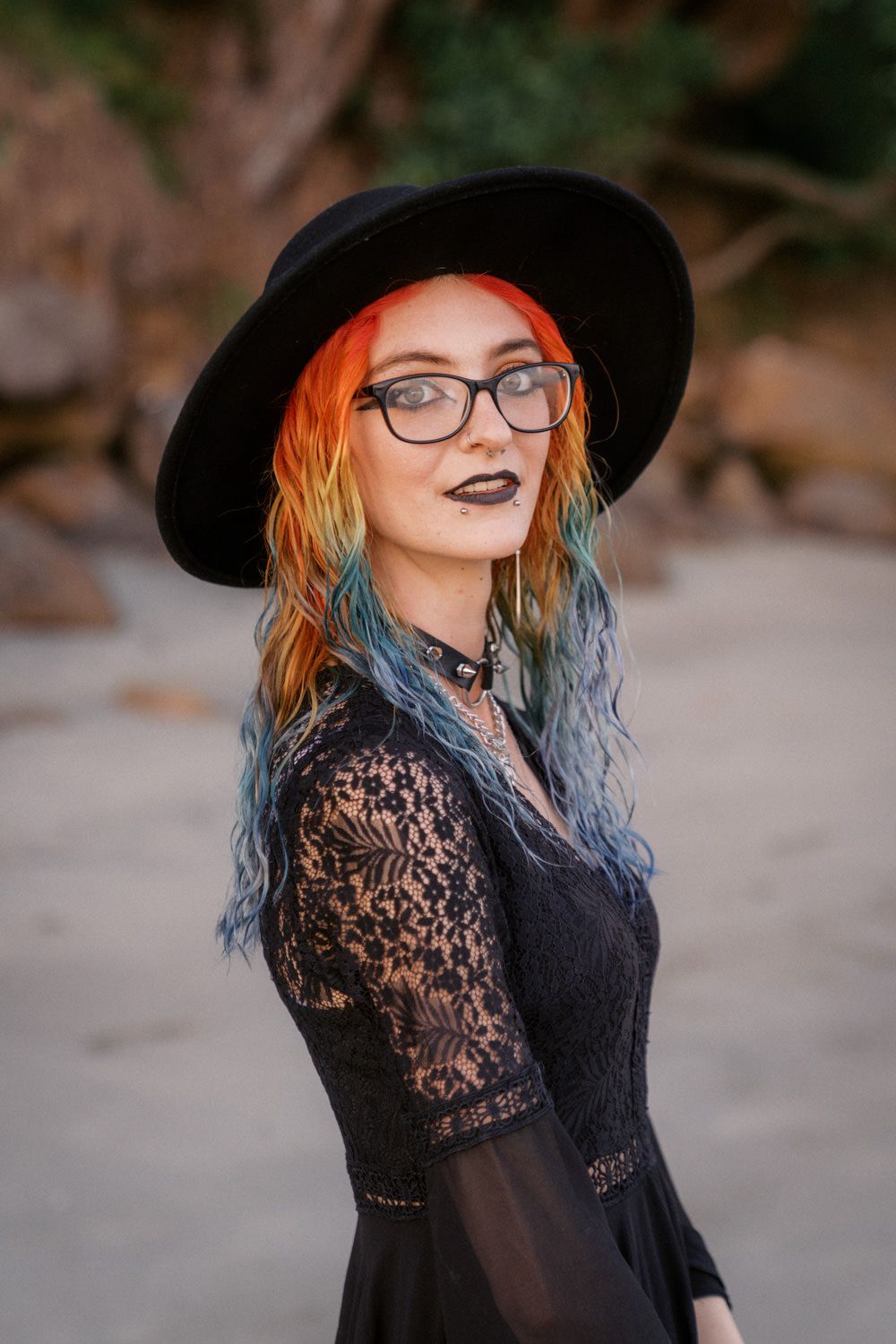 Woman with rainbow hair wearing black lace dress, wide-brim hat, and glasses poses outdoors.