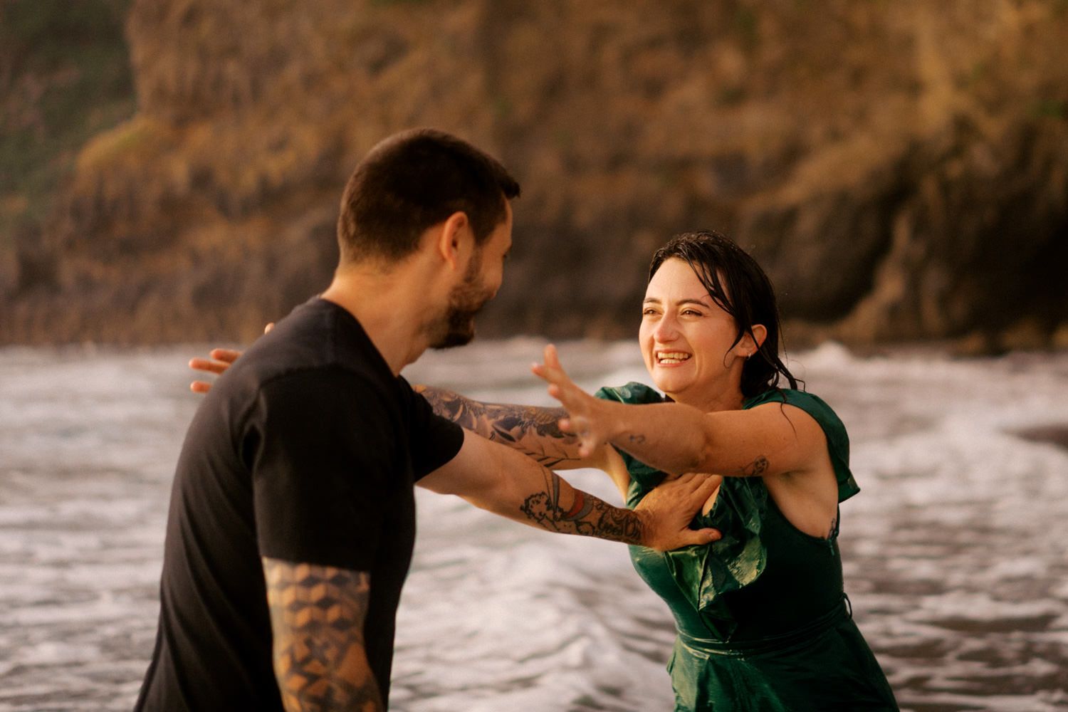 A smiling couple playfully dancing together on a rocky beach, the woman wearing a green dress and the man with tattoos.