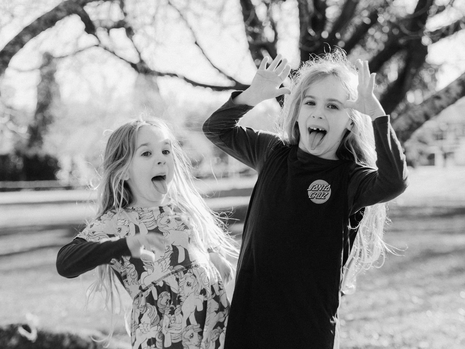 Two young girls make silly faces and stick out their tongues in a fun black-and-white outdoor photo.