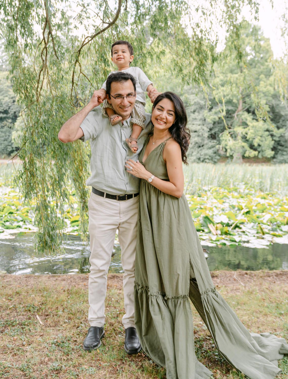 Family of three posing outdoors near a pond with willow trees, parents smiling as child sits on father's shoulders.