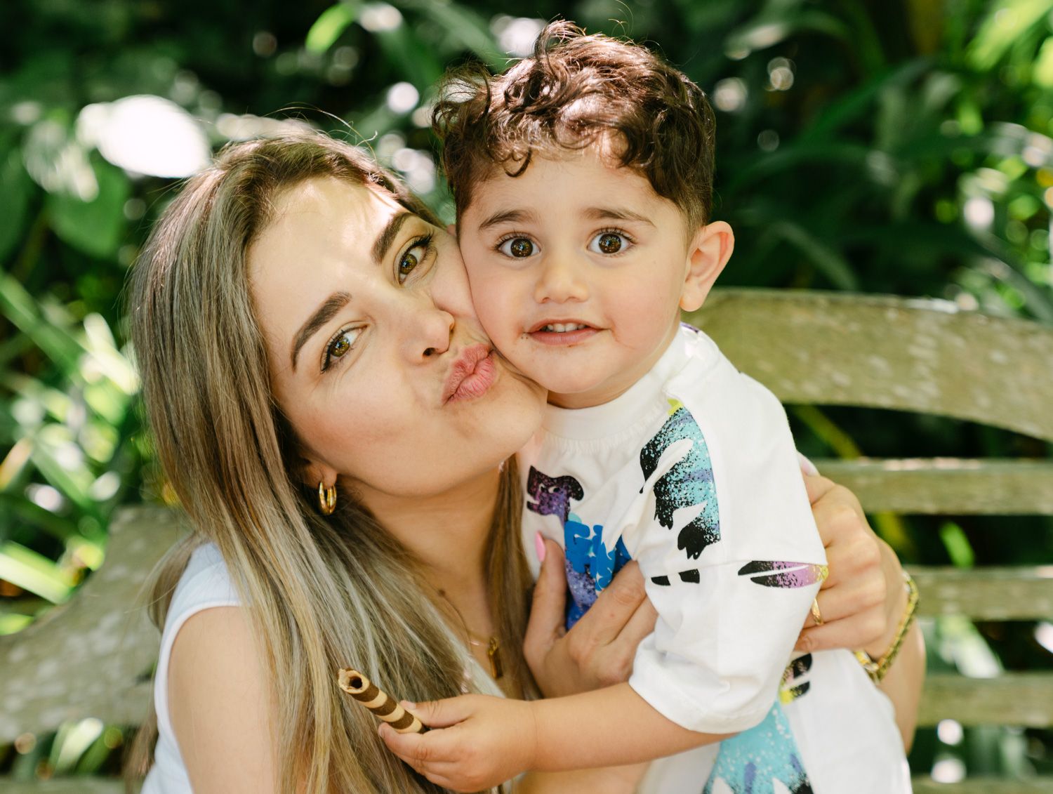 A young woman kisses a curly-haired toddler boy outdoors among lush green foliage.