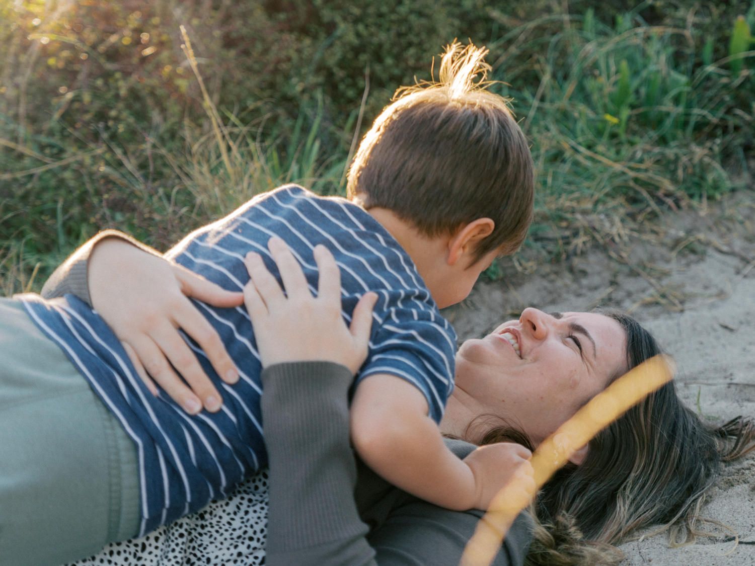 A mother lying on sand smiling at her young son who sits on her lap outdoors in nature.
