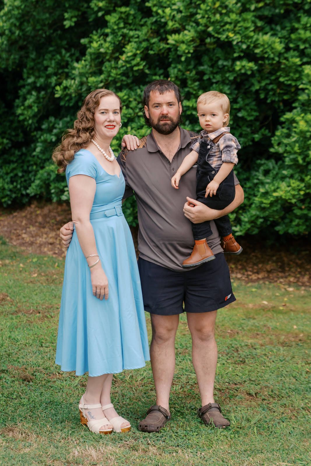 Family portrait of a woman in a blue dress, a bearded man, and a toddler boy posing on green grass outdoors.