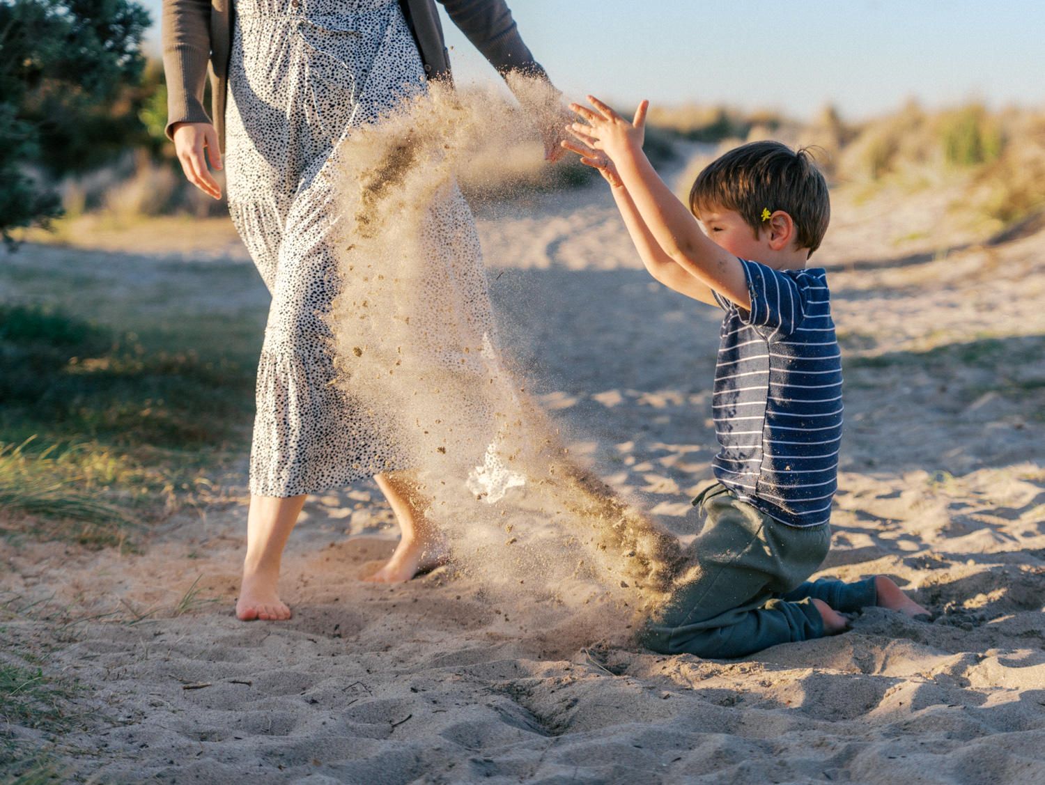 A young boy sits on sandy beach throwing sand while a woman in floral dress stands nearby on a sunny day.