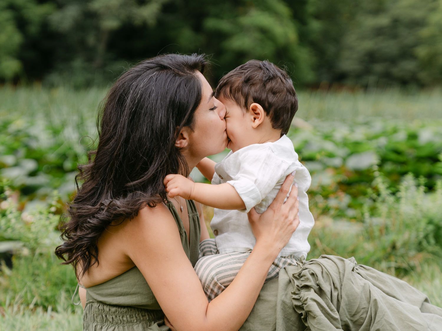 A mother tenderly kisses her baby boy outdoors in a lush green garden setting.