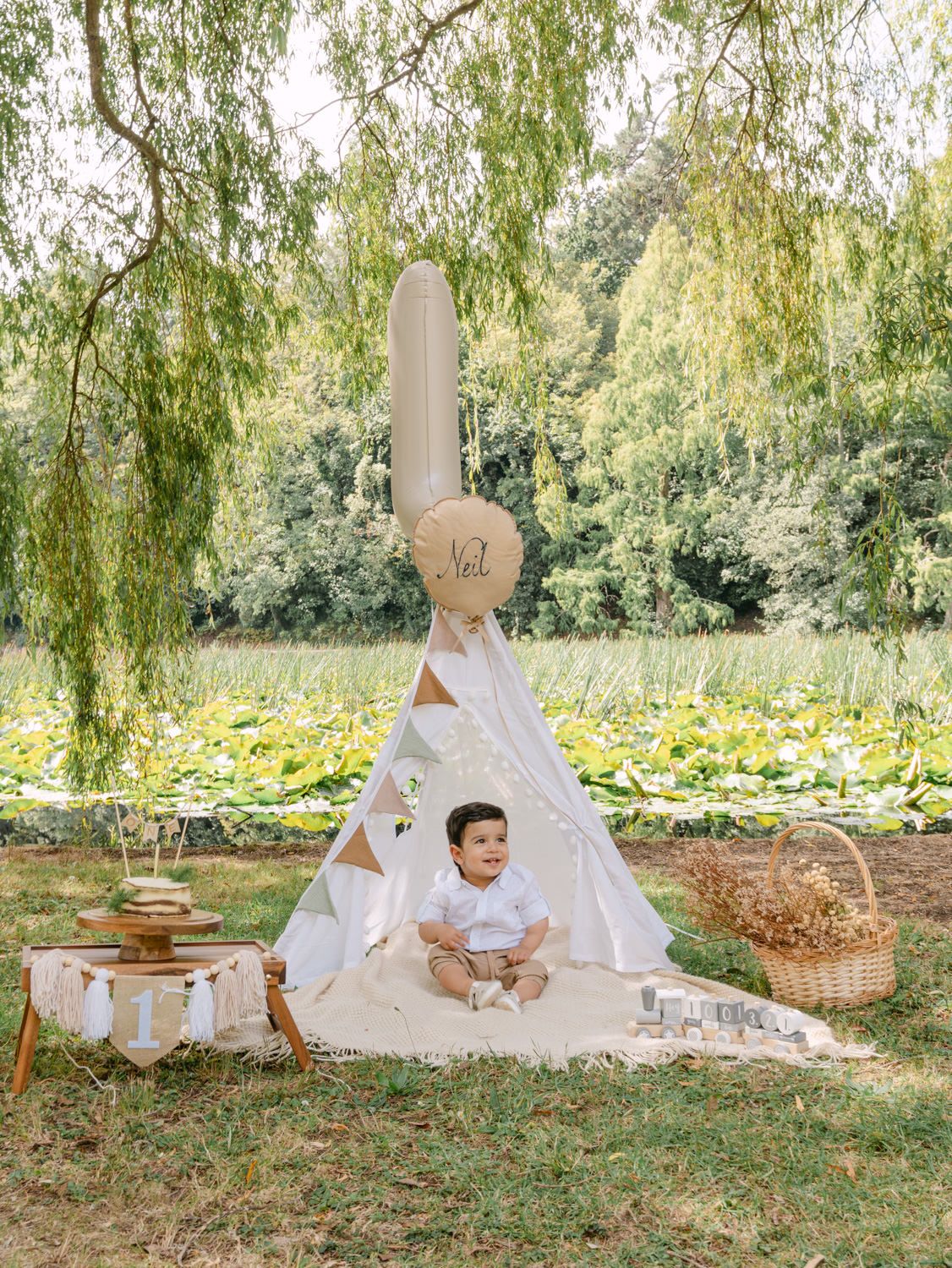 A baby boy sits in front of a white teepee tent on a blanket outdoors, surrounded by birthday decorations near a pond.